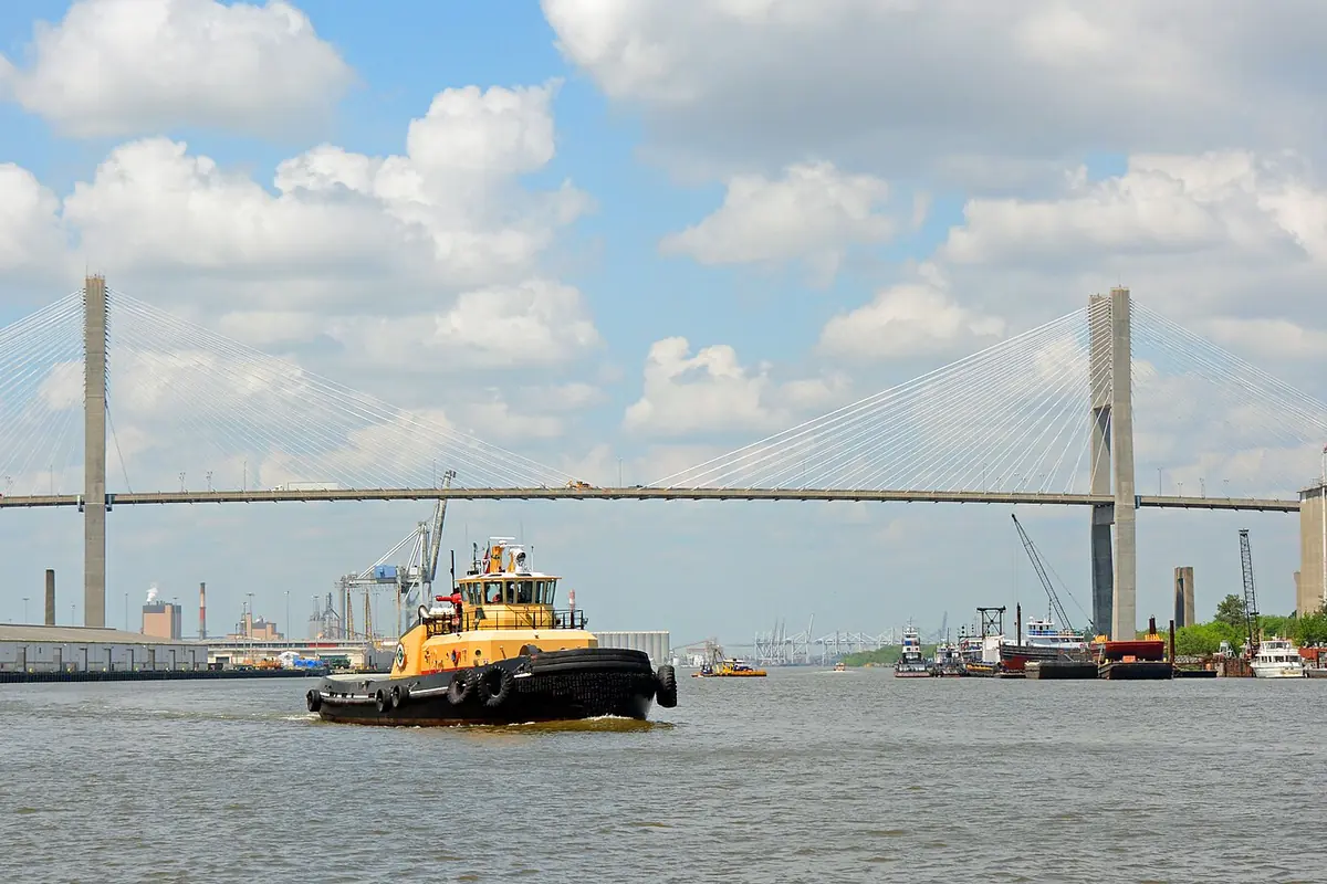 흐린 푸른 하늘 아래, 쌍둥이 탑이 있는 Talmadge Memorial Bridge 앞 Savannah River 위를 지나는 노란색과 검은색 예인선.